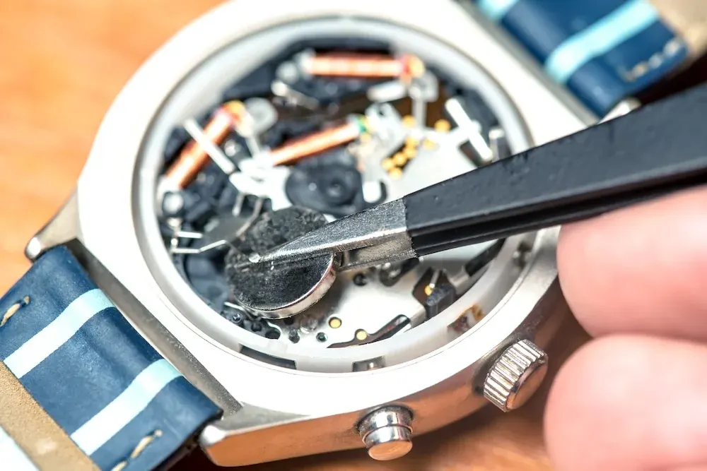 A jeweler replacing a watch battery with tweezers inside an open watch case.